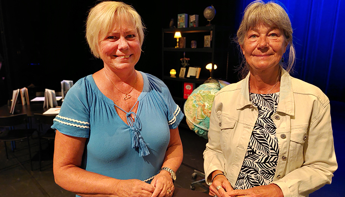 Två kvinnor står vid ett runt svart bord. De har händerna på bordet och ser glada ut.