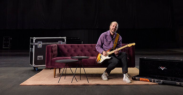Man sitter på en stor svart låda. Han håller i en gitarr